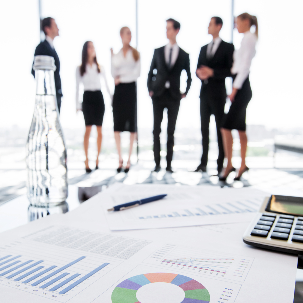 Immagine di un gruppo di professionisti in abbigliamento formale che discute in ufficio, con documenti finanziari, grafici e una calcolatrice in primo piano. La scena rappresenta la pianificazione aziendale e l'importanza di mantenere assetti organizzativi adeguati per una gestione efficace.